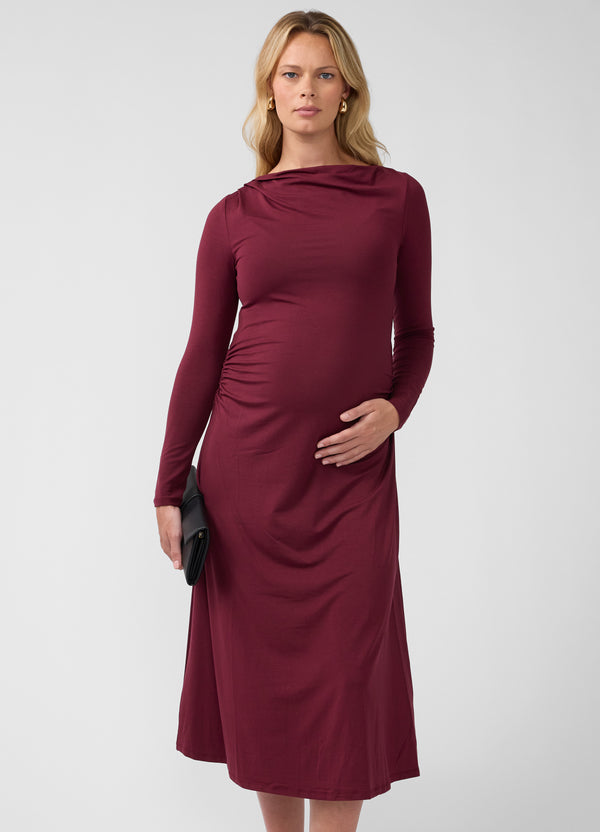 A pregnant woman wearing a burgundy ruched long sleeve mock neck maternity midi dress while holding a black clutch