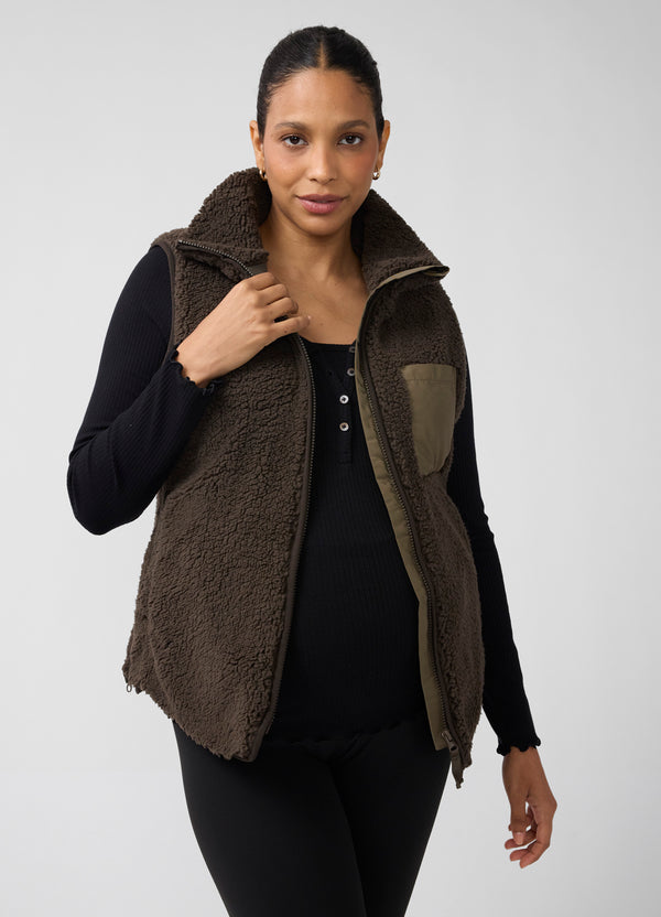 A pregnant woman modeling a brown sherpa fleece maternity vest over a black long-sleeve top and black leggings, photographed against a light gray studio background.