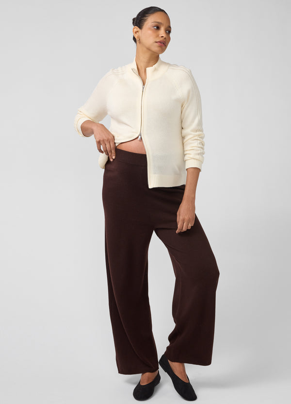 A pregnant woman wearing a dark brown wide leg knit maternity sweater pants with an off-white double-zip ribbed knit cardigan and black ballet flats