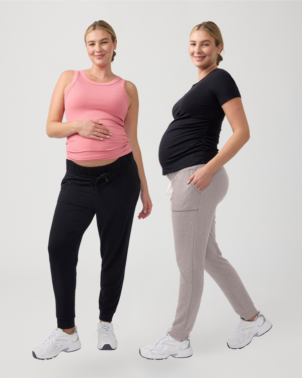 ||2 pregnant women, one wearing a pink tank top and black knit maternity jogger pants, and the other wearing a black t-shirt and stone grey knit maternity jogger; both in white sneakers