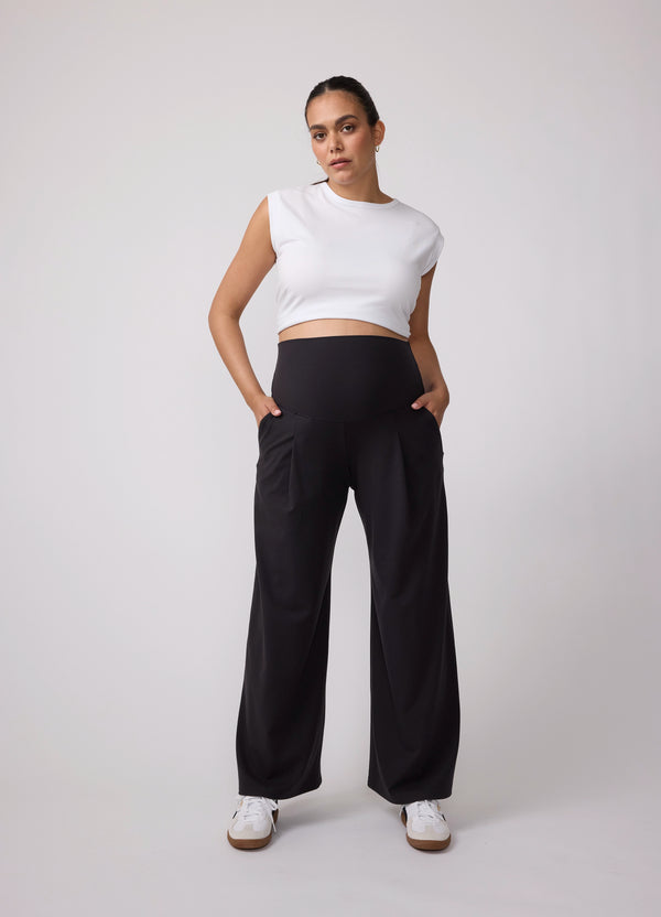 A pregnant woman wearing a black active and everyday maternity wide leg pants with a white cropped shirt and white sneakers
