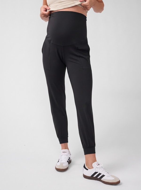 A half body shot of a pregnant woman wearing a black active tapered leg knit maternity joggers with white sneakers