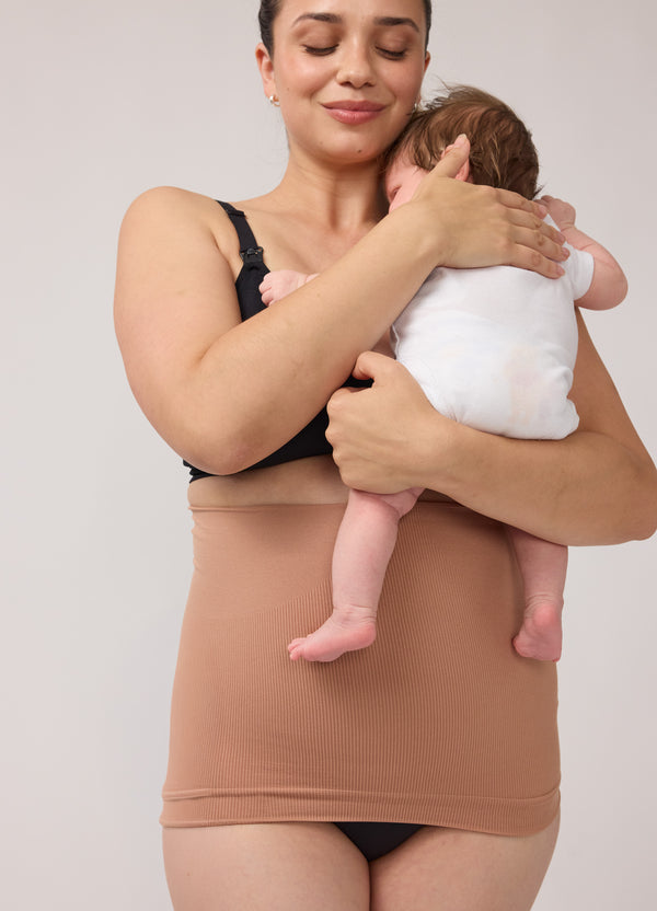 A half body shot of a woman wearing a black bra and underwear with a mocha-colored afterband belly and back postpartum support band while holding a baby
