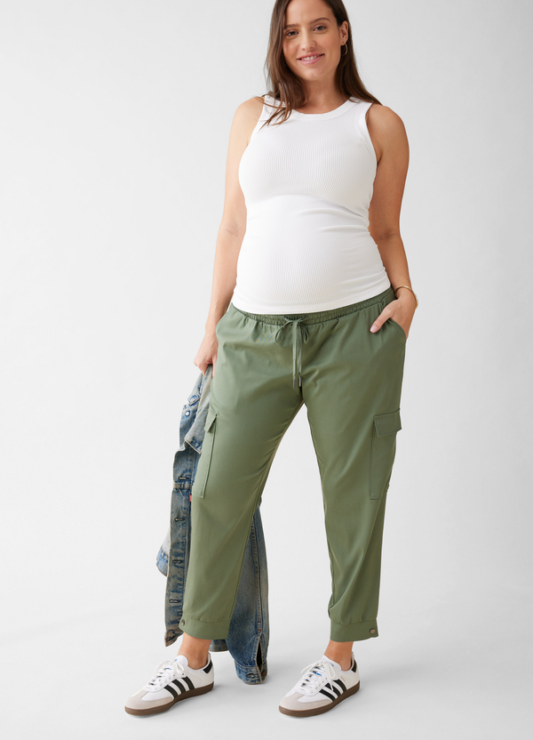 ||A pregnant woman holding a denim jacket on her right hand while wearing green cargo maternity jogger pants and a white ribbed tank top, with white sneakers 
