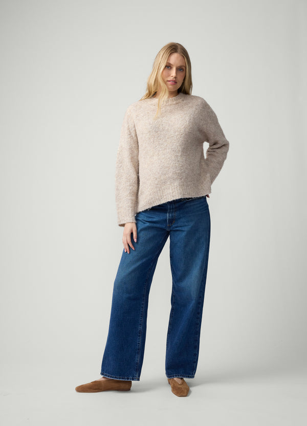 A pregnant woman wearing a beige cosy knit sweater, dark wash denim jeans, and brown suede ballet flats