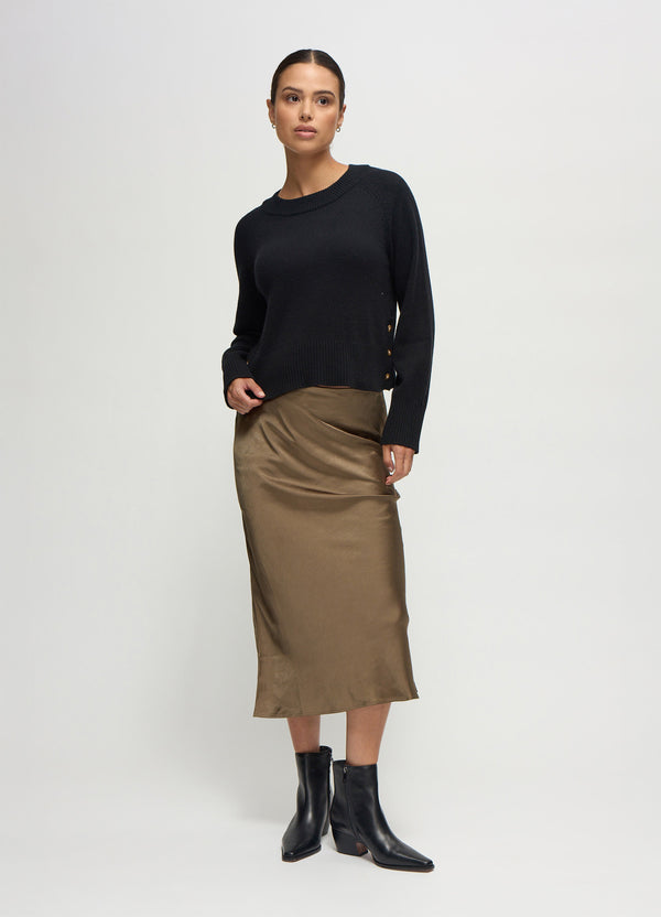 ||A pregnant woman wearing a black maternity sweater, an olive satin slip skirt, and black ankle boots