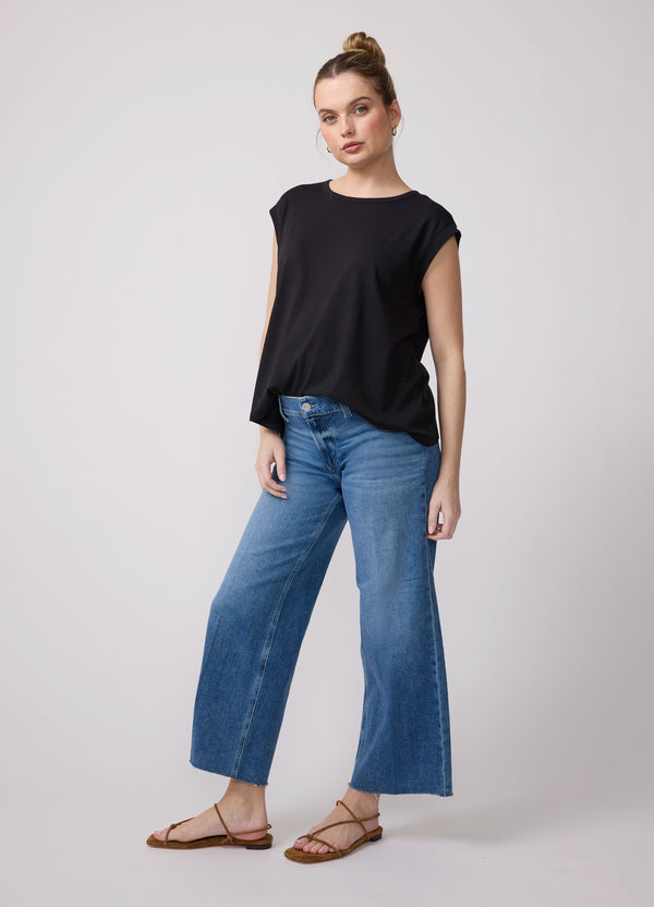 A pregnant woman wearing a black relaxed crew neck cotton maternity tank with blue denim jeans and brown strappy flat sandals