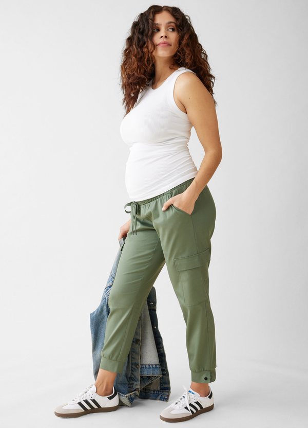 A side view shot of a pregnant woman wearing a white seamless ribbed high neck maternity tank top with olive green cargo jeans and white sneakers, while holding a denim jacket