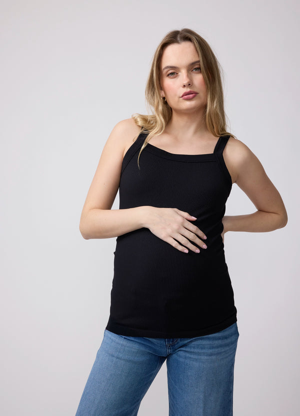 ||A half body shot of a pregnant woman with one hand on her hip and the other on her belly, while wearing a black seamless ribbed maternity tank top with blue straight denim jeans