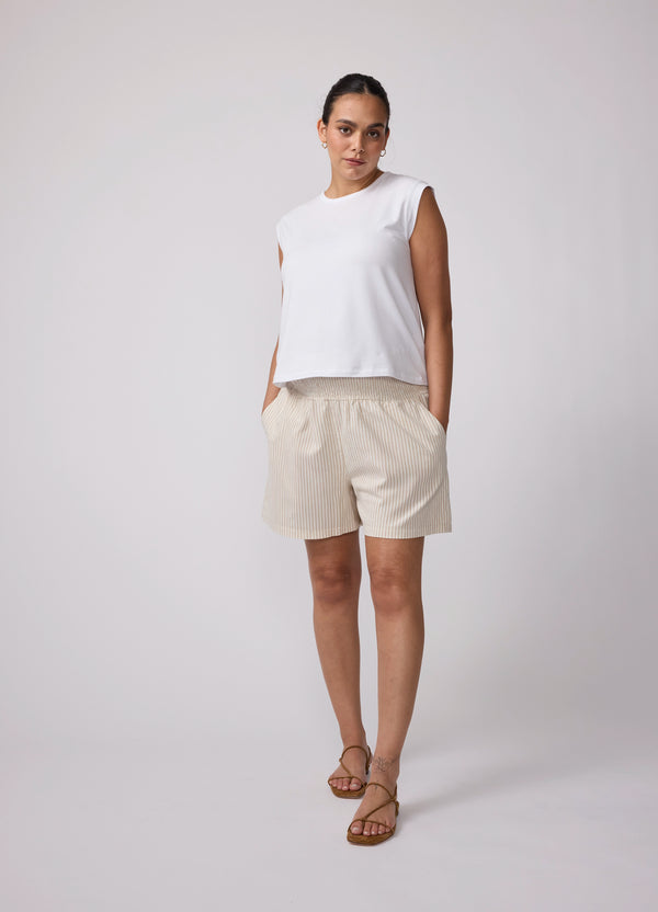 A pregnant woman with her hands in the pockets of the tan and white smocked wide leg striped linen maternity shorts she's wearing with a white muscle tee and a pair of brown strappy flat sandals