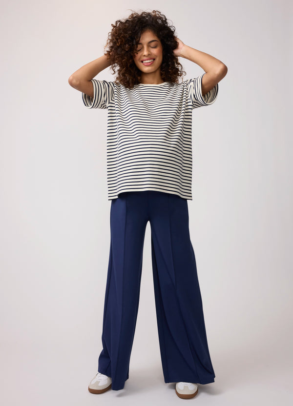 A pregnant woman sitting on the floor while wearing a navy blue and white striped weekend crew maternity sweatshirt with blue pants, and white sneakers