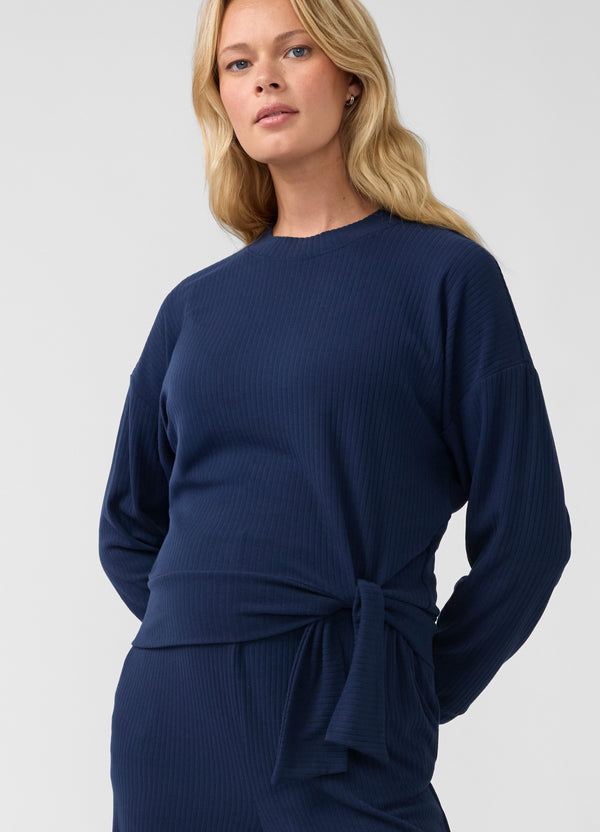 A pregnant woman wearing a navy blue ribbed knit long-sleeve maternity top with a self-tie waist belt, shown against a plain light gray studio background.