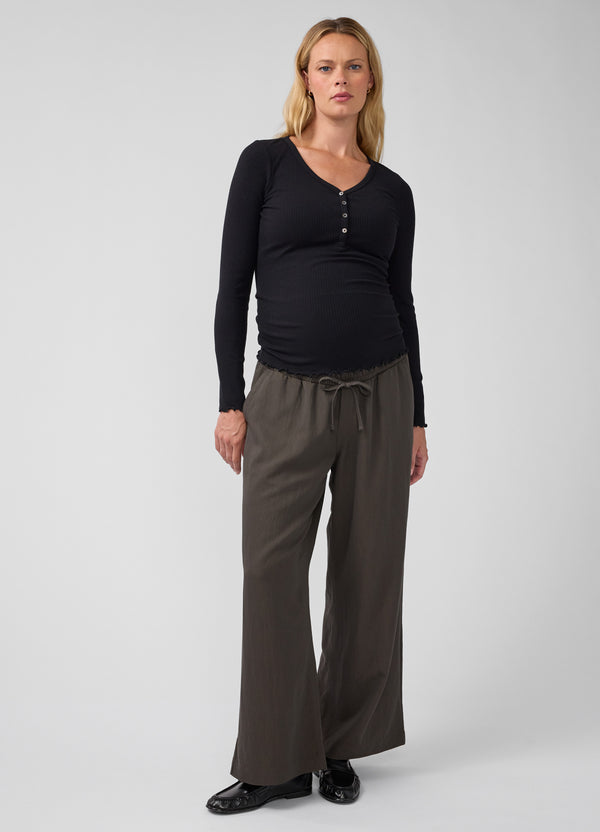 A pregnant woman modeling a black long-sleeve henley maternity top and dark olive green wide-leg maternity pants against a light gray studio background.