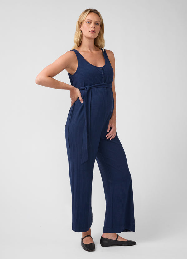 A pregnant woman modeling a navy blue maternity jumpsuit with a tie waist and wide legs, paired with black flats, against a light gray studio background.