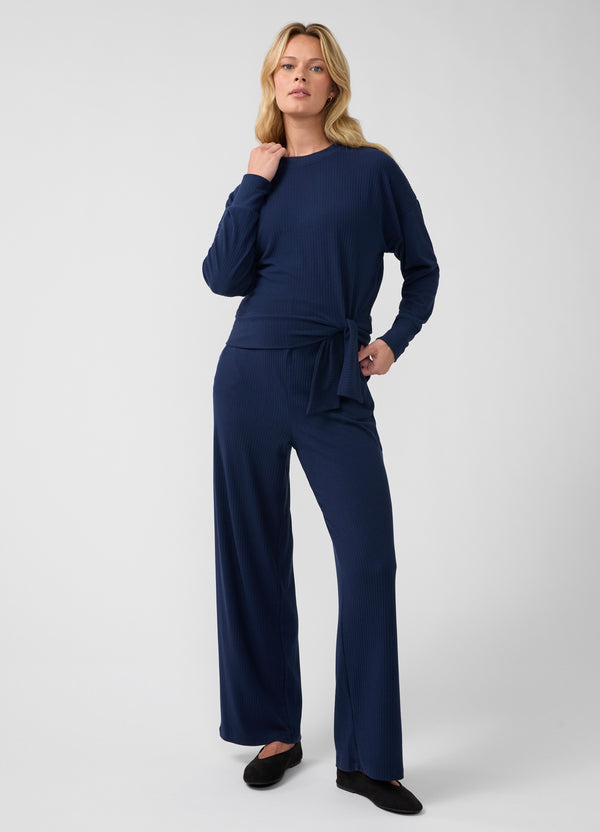 A pregnant woman modeling a navy blue long-sleeve maternity jumpsuit with a tied waist and wide-leg maternity pants, standing against a light gray studio background.