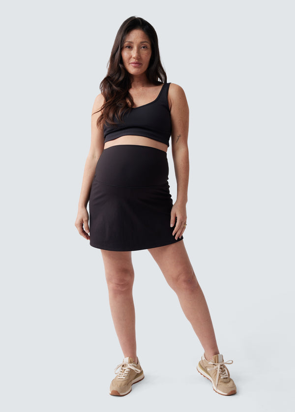 ||Pregnant woman wearing a black active skirt, a black sports bra, and beige sneakers