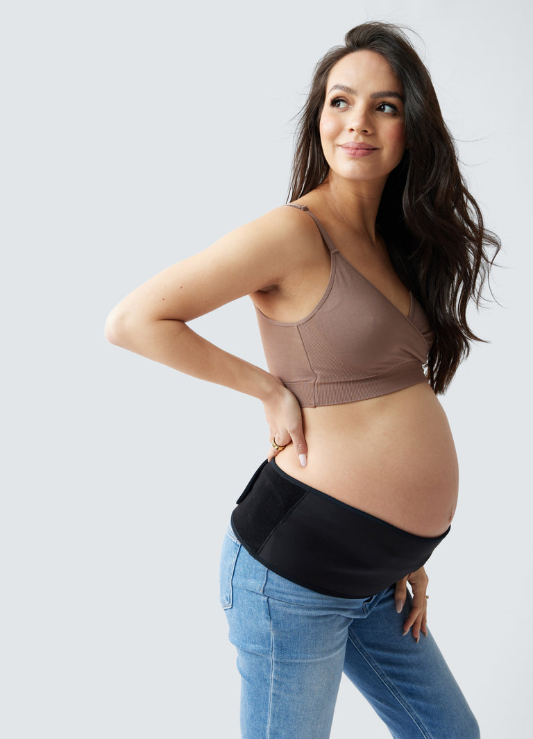 ||A pregnant woman wearing taupe bra, black pregnancy support belt, and blue jeans on a plain background 