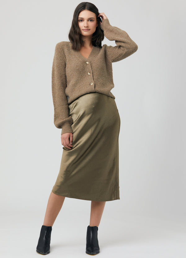 A pregnant woman models an olive green satin maternity midi skirt with a beige knit cardigan and black boots.